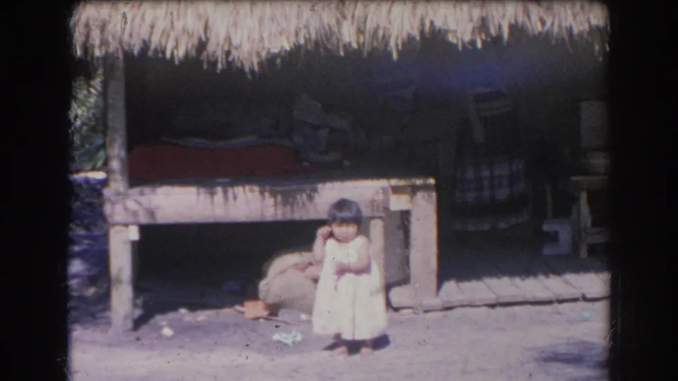 A child in a light dress stands before a rustic thatched structure with shaded seating and wooden posts visible behind.