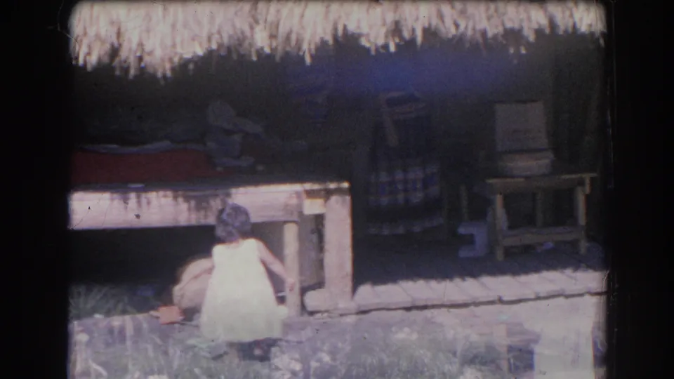 The child turns away from the camera near the thatched-roof structure, with benches and shaded objects in the background.