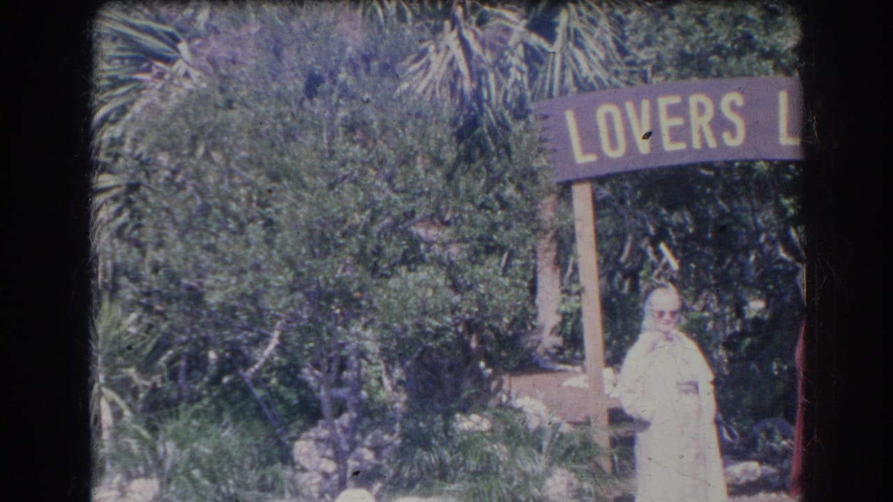 Older woman in a light coat standing near a Lovers Lane sign in a leafy garden.