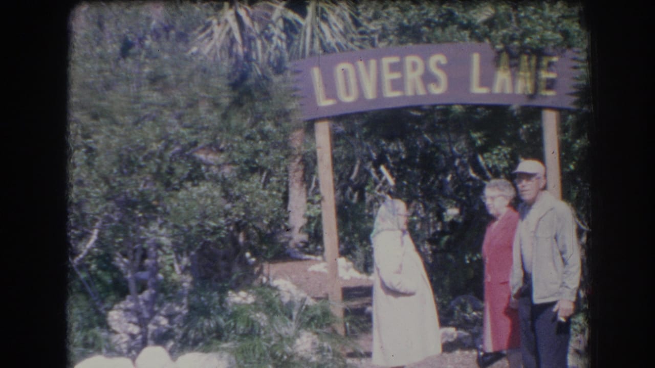 Three older adults standing together beside the Lovers Lane sign and garden plants.