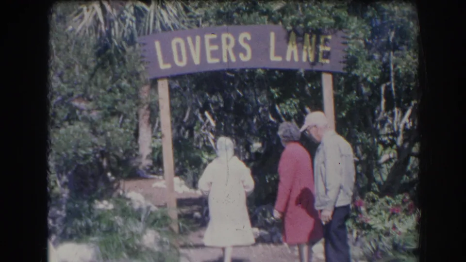 1969 Sarasota 8mm frame showing older adults clustered near the Lovers Lane sign, with foliage and flowers around them.