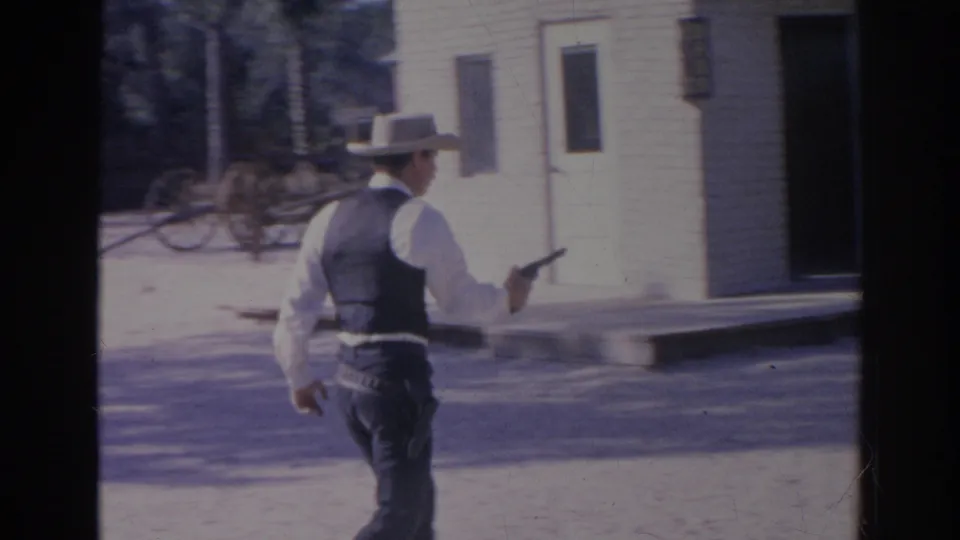 A cowboy-hatted man in vest and white shirt walks forward holding a handgun in a staged Western setting.