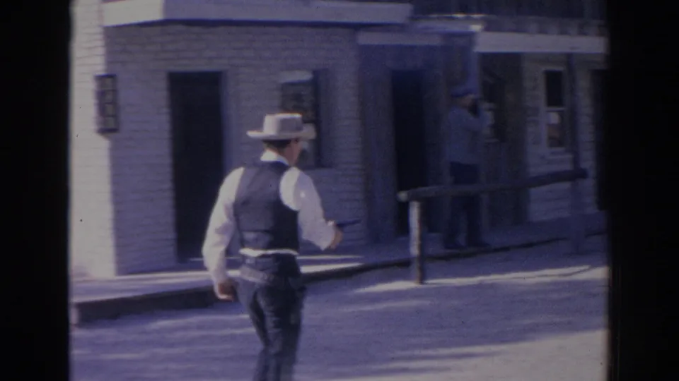 A cowboy-hatted man in vest points a handgun toward a person near a Western-style building in 1969 home-movie footage.