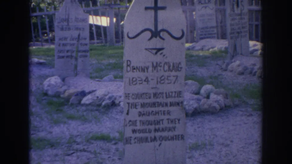 1969 Sarasota 8mm frame showing a wooden Benny McChang grave marker, a cross-shaped top, surrounding stones, grass, and other markers behind it.