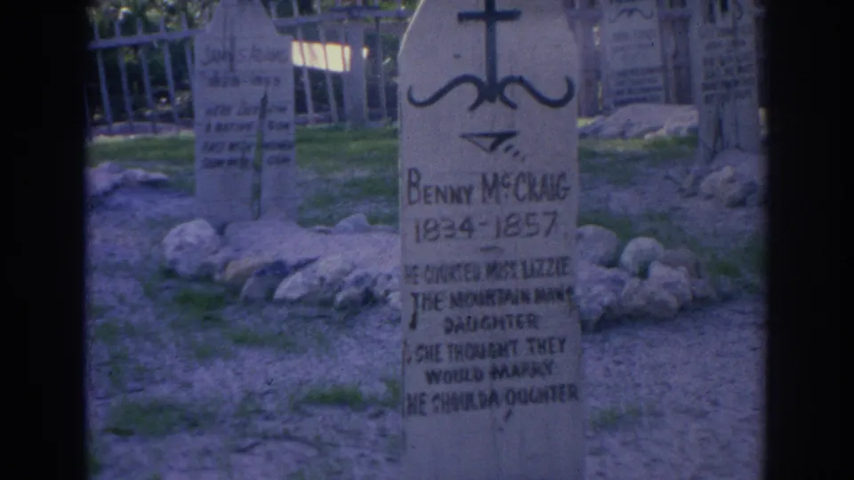 Centered 1969 Sarasota frame of the Benny McChang wooden marker, with painted epitaph lines, rocks at the base, grass, and background markers.