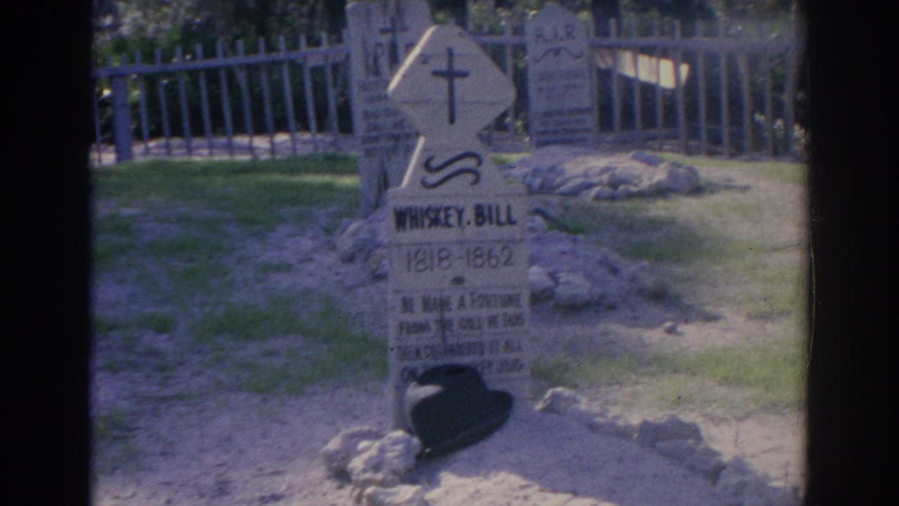 Cemetery headstone with cross symbol, low stones, grass, and pale fence in sunlight.