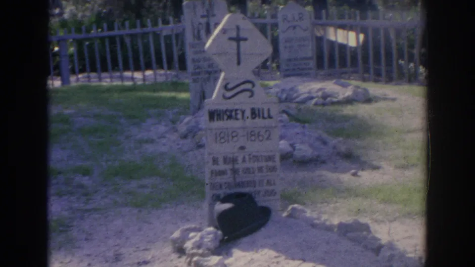 1969 Sarasota home-movie frame showing a weathered cemetery headstone, cross motif, low stones, grass, and a pale fence.