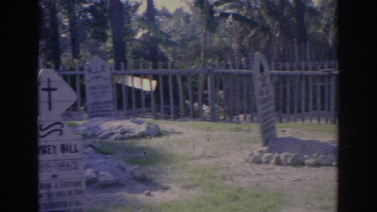 1969 Sarasota cemetery view with multiple headstones, a pale fence, grass, trees, and handheld 8mm framing.
