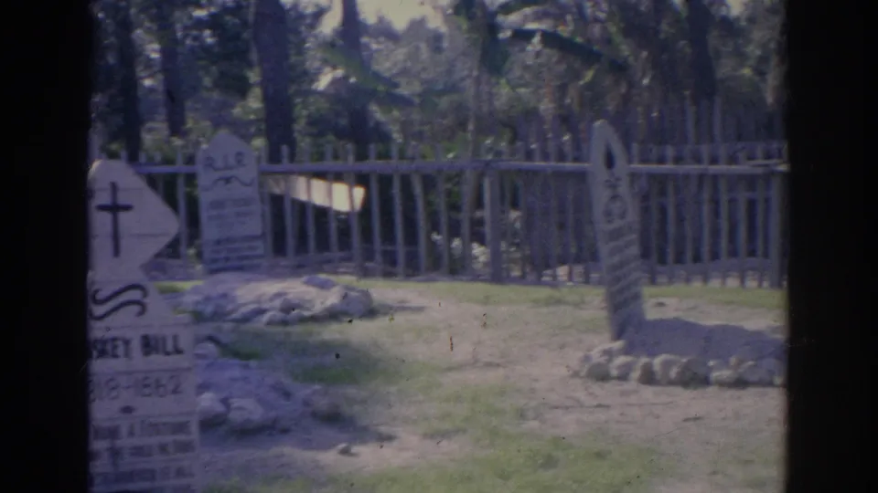 1969 Sarasota cemetery view with multiple headstones, a pale fence, grass, trees, and handheld 8mm framing.