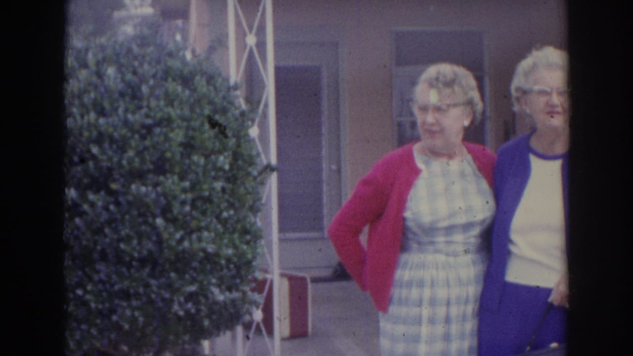 1969 Sarasota home-movie frame showing two women posing outside a building with shrubs, porch posts, windows, and luggage nearby.