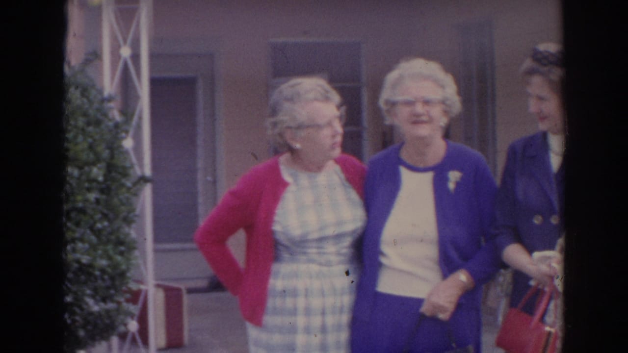 1969 Sarasota home-movie frame showing three women outside a building, including one holding a red handbag, with shrubs and windows behind them.