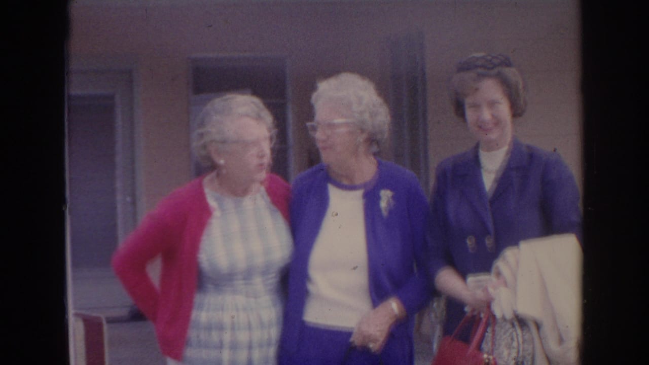 Three older women standing together outside a building, with one in a red cardigan and two in blue outerwear.