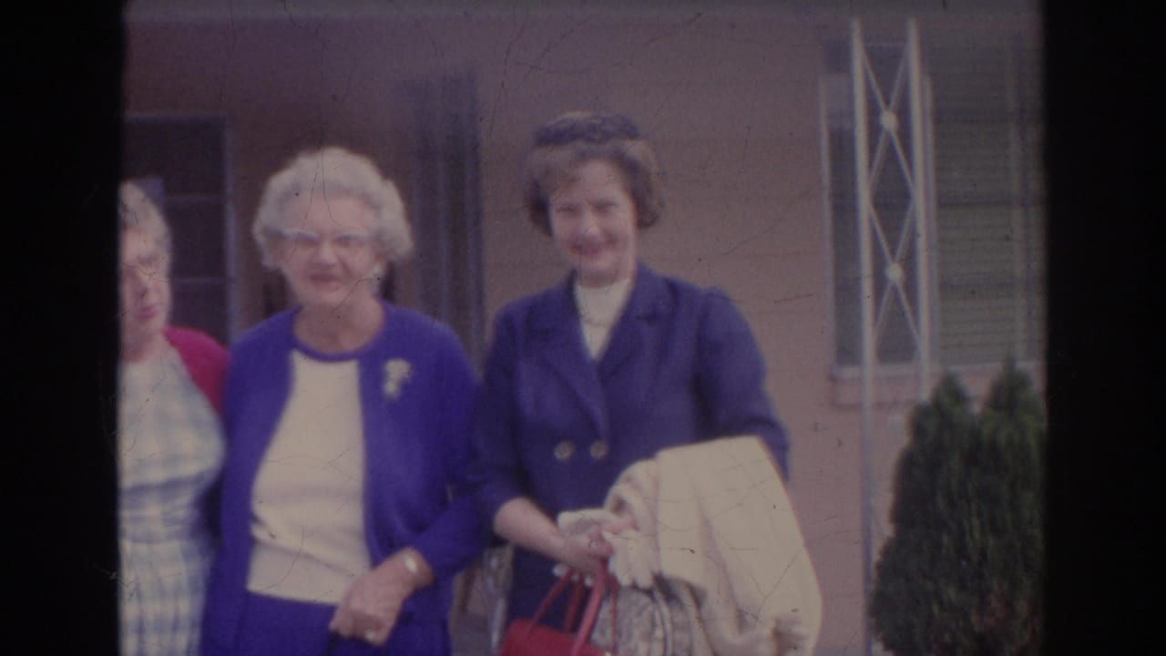 Three older women smiling near a building, with a partial person at the left edge and porch detail behind them.