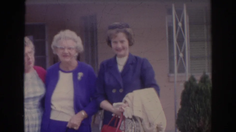Three older women smile toward the camera in Sarasota, Florida, with a building facade and porch detail behind them on 8mm film.