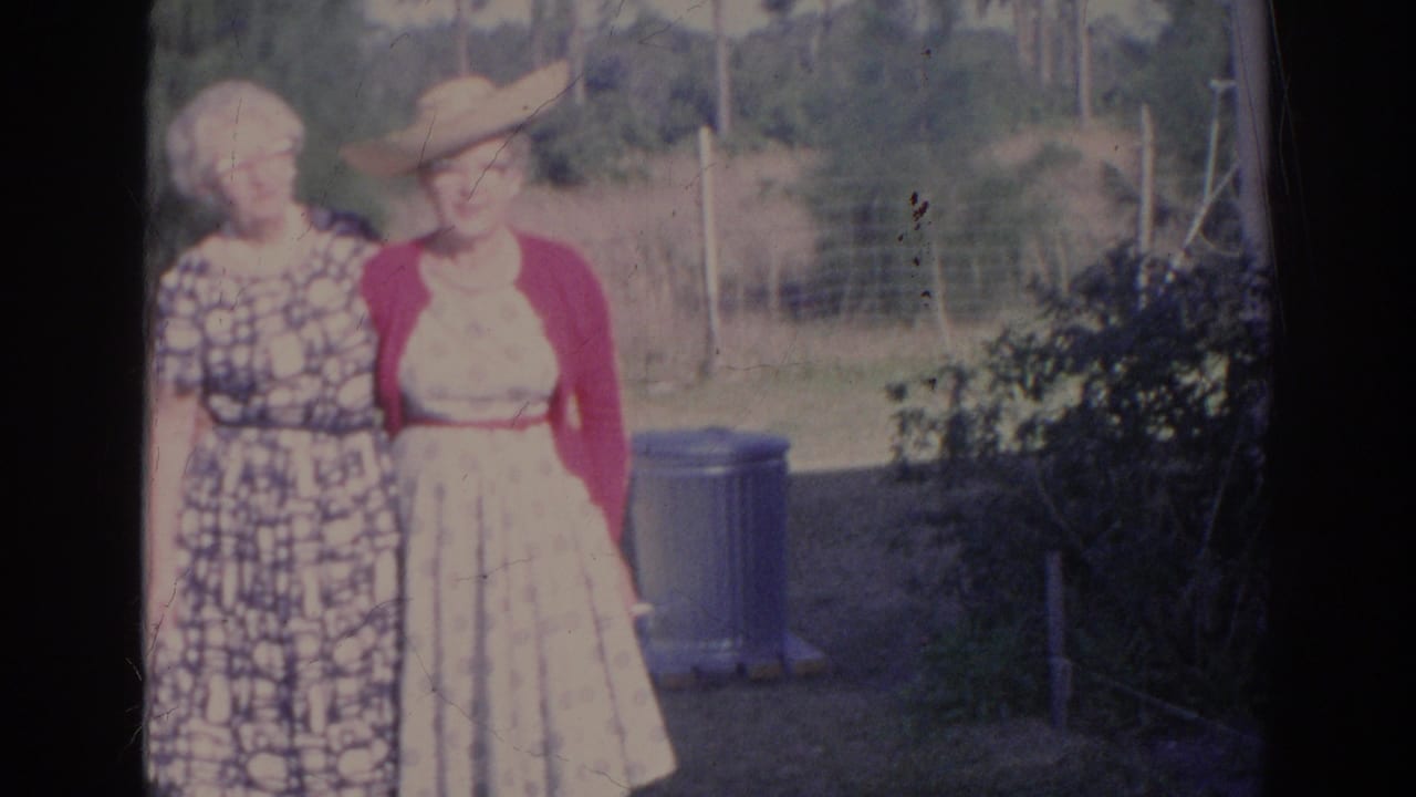 Two elderly women stand together in a sunny yard near trees and a blue trash can.