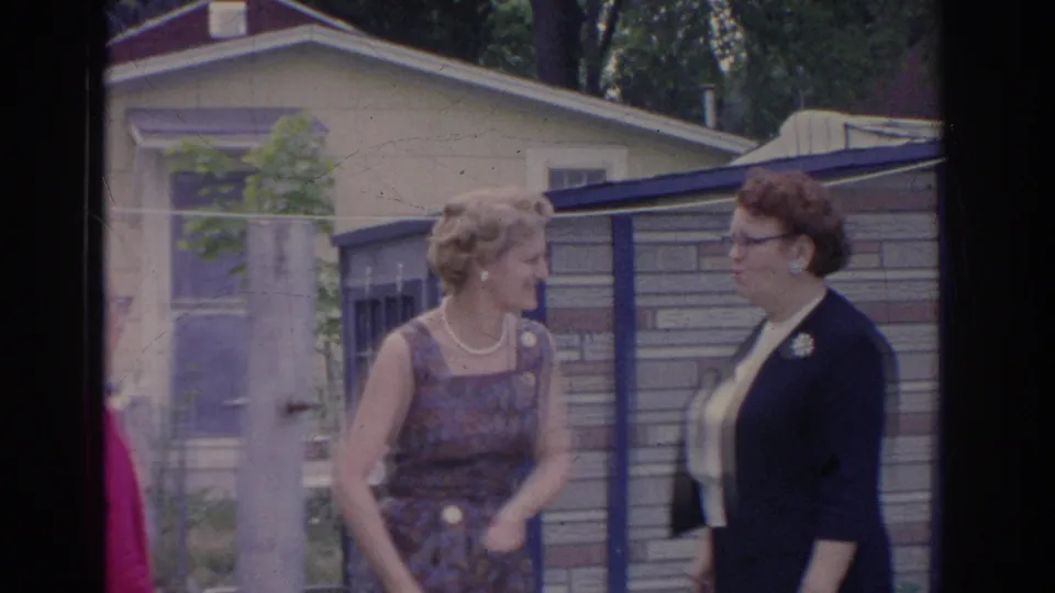 In 1969 Sarasota home-movie footage, two women converse beside a fence and nearby house in a suburban yard.