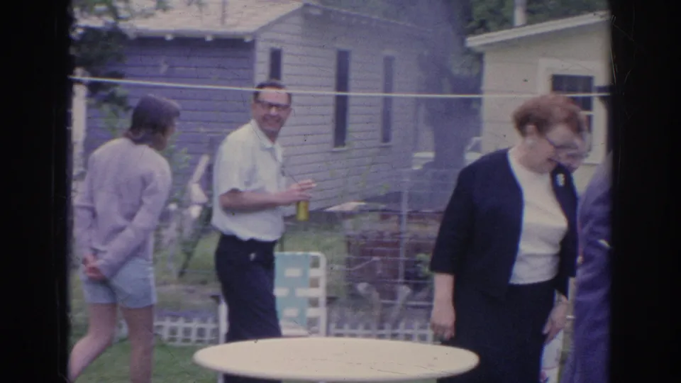 Adults gather in a 1969 Sarasota suburban yard, with a round table, fencing, trees, and house exteriors visible.