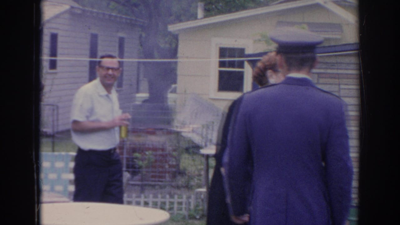 Man in a light shirt stands outdoors as a uniformed man faces a small group.