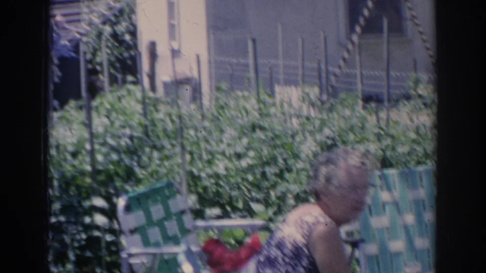A woman sits or leans near green lawn chairs and garden plants in the Sarasota backyard, with fencing and a building behind her.