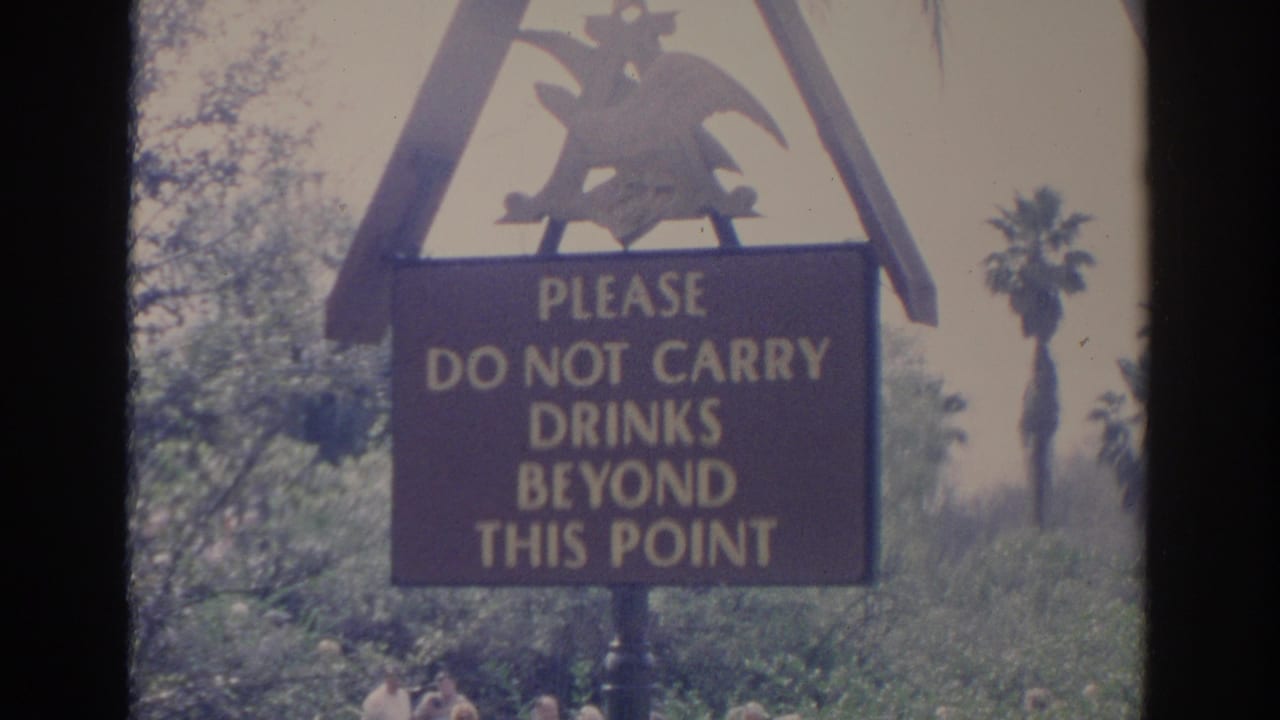 Drink-warning sign beside palm trees and greenery near a water channel.
