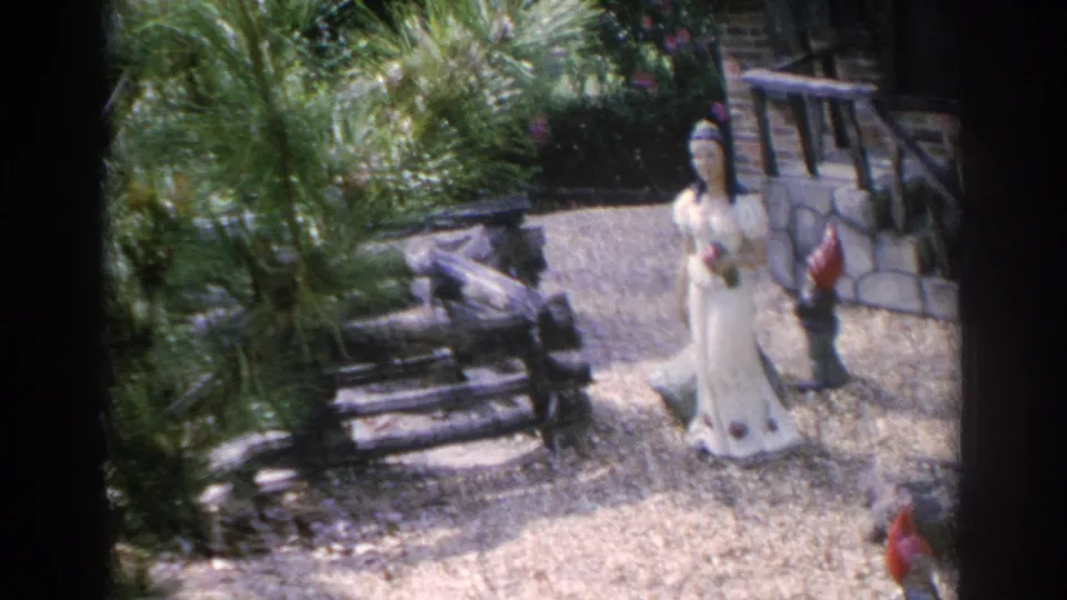 1969 Sarasota 8mm view of a storybook garden display with a white figure, dwarf statues, foliage, and stonework.