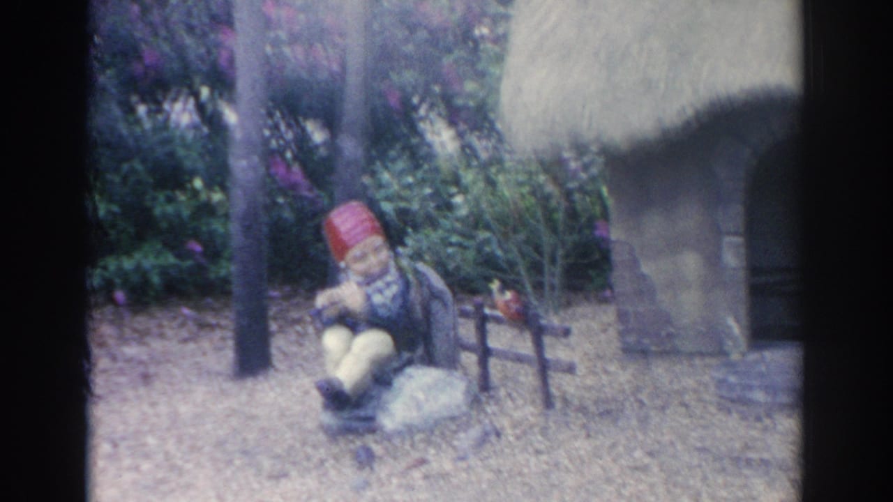 1969 Sarasota 8mm frame showing a red-hatted gnome figure near a bench and miniature cottage in a leafy garden.