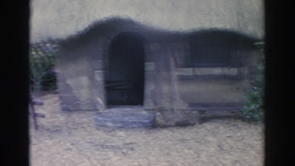 1969 Sarasota home-movie frame centered on a miniature thatched cottage with dark doors, a small window, and garden planting.