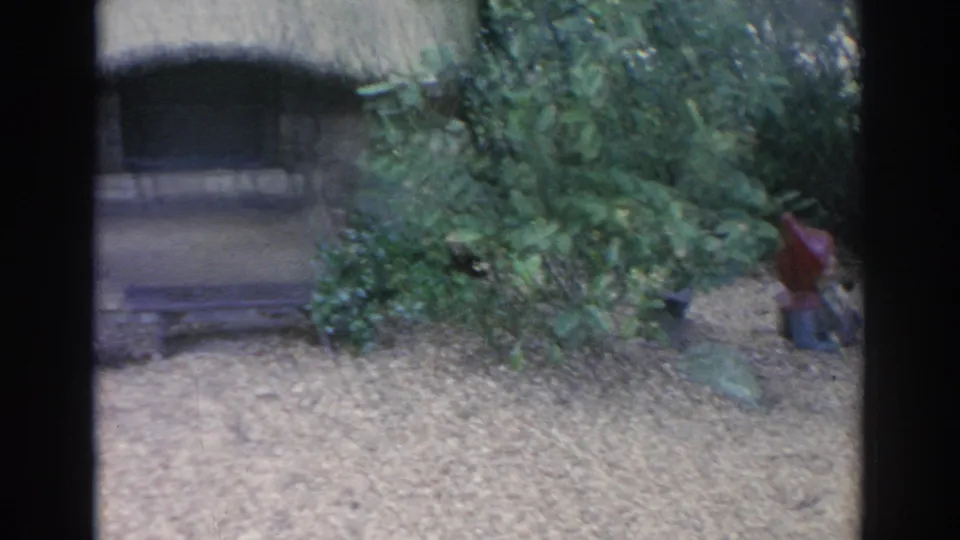 1969 Sarasota 8mm frame showing the cottage edge, dense greenery, and a small red-hatted figure near the garden border.