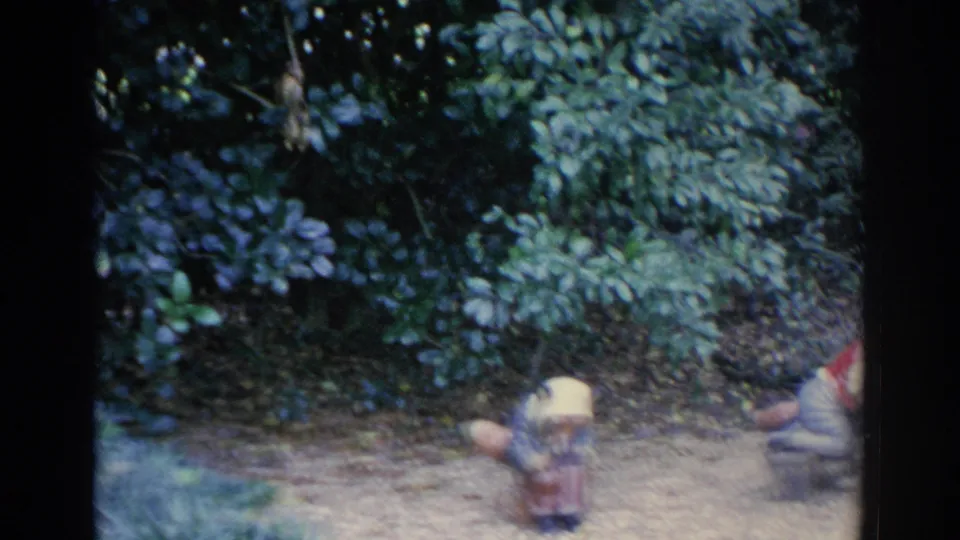 1969 Sarasota home-movie frame showing dense shrubs, a small yellow-hatted garden figure, and part of a seated red-hatted figure.