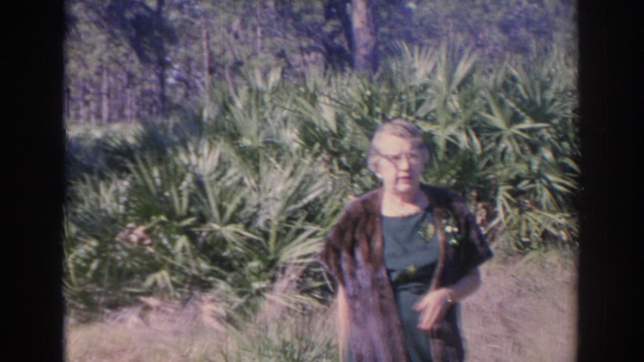 Older woman facing the camera beside dense palm fronds in bright sunlight.