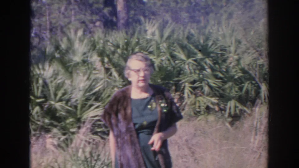 Older woman in a dark dress and brown wrap stands amid palms and dry grasses in Sarasota, Florida, captured on 1969 8mm film.