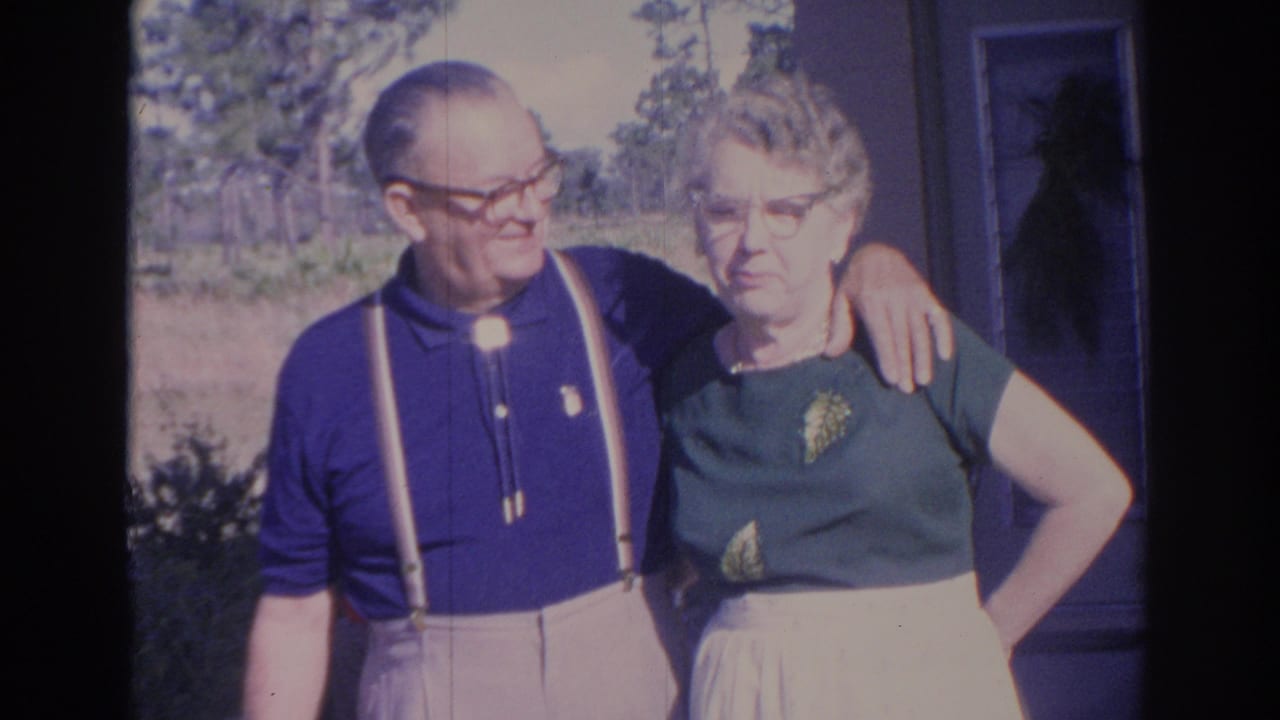 In metadata-supported 1969 Sarasota footage, an older couple stands together outdoors beside a house with shrubs and trees behind them.