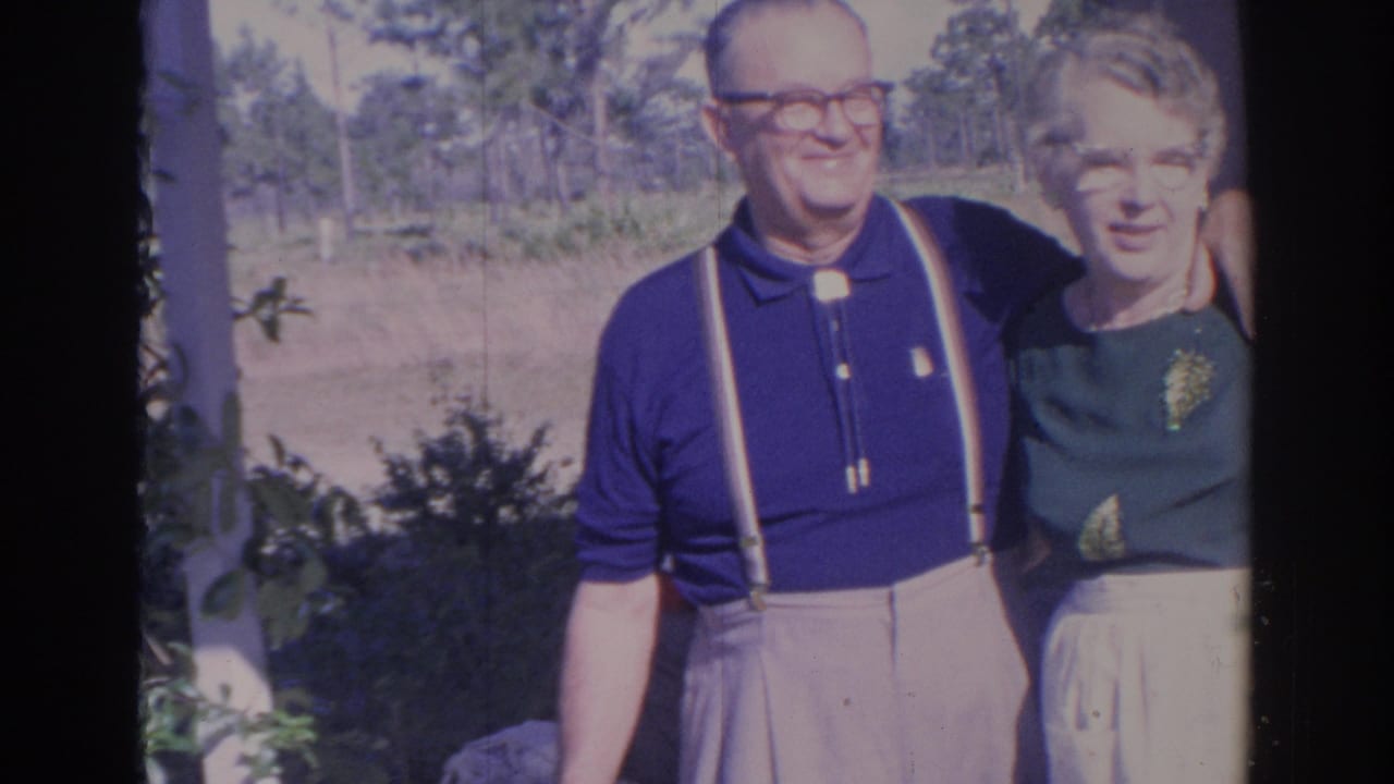 Older couple facing the camera near shrubs, with trees and bright yard behind them.