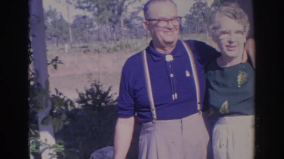 The couple remains posed outdoors in Sarasota, with the man in suspenders and the woman in a green top near sunlit greenery.