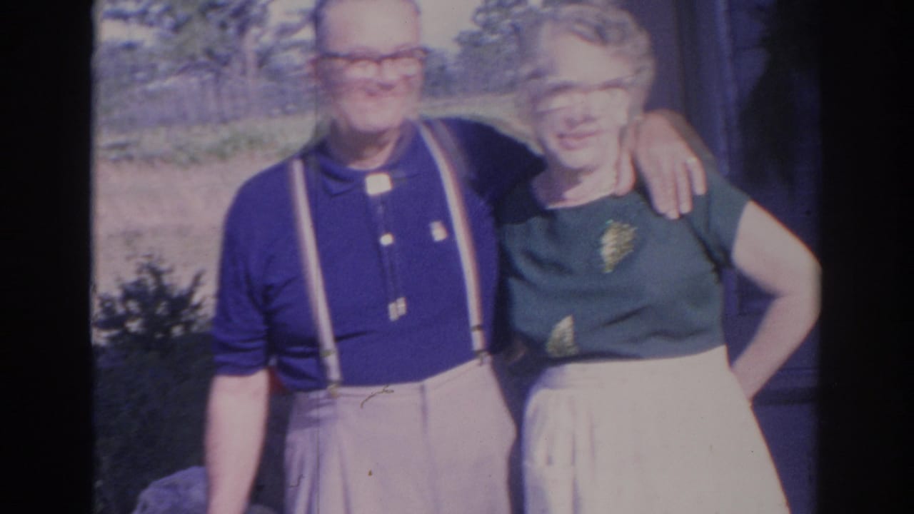 A slightly blurred 8mm frame shows the older couple standing arm in arm outside, with the house edge and yard visible behind them.