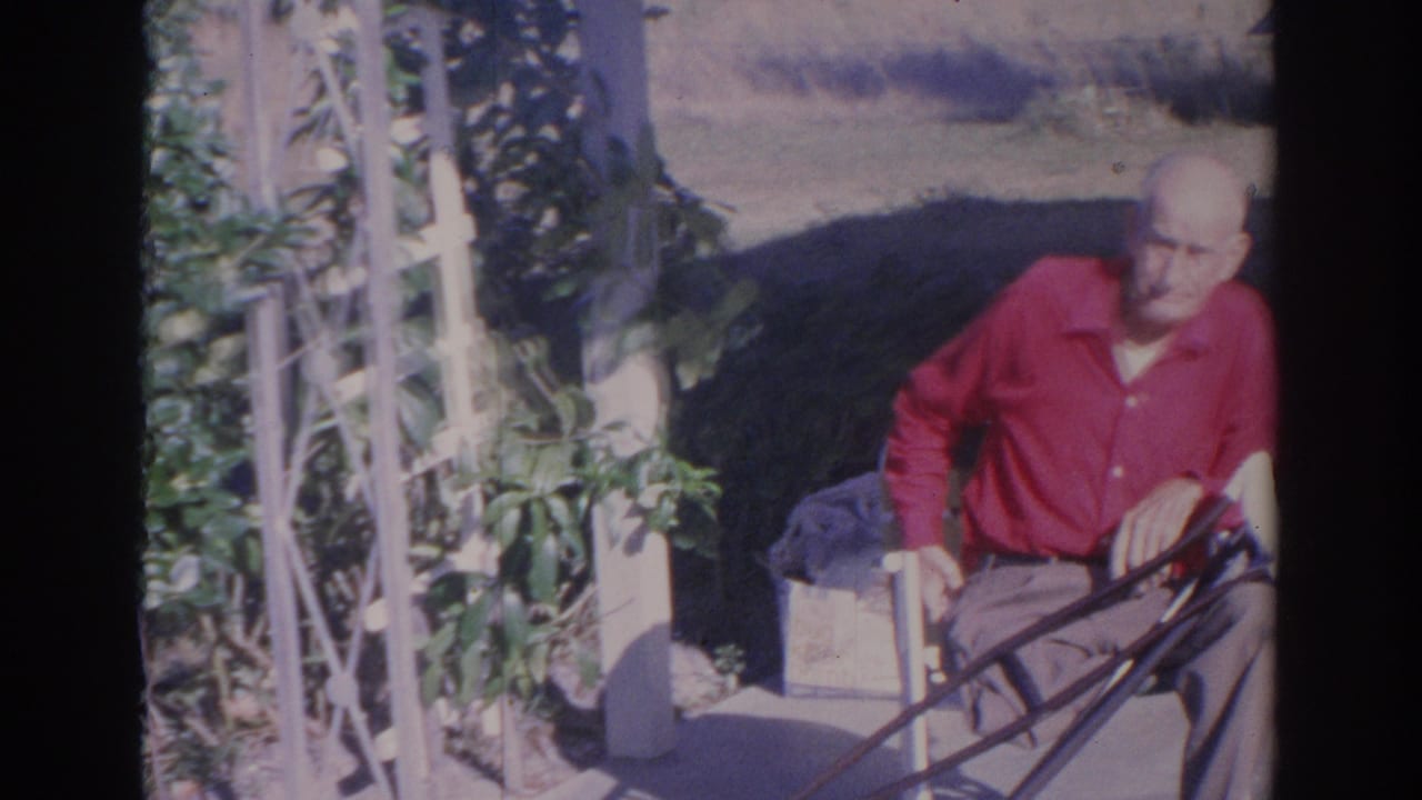 Older man in a red shirt sits in a white outdoor chair beside a trellis and greenery.