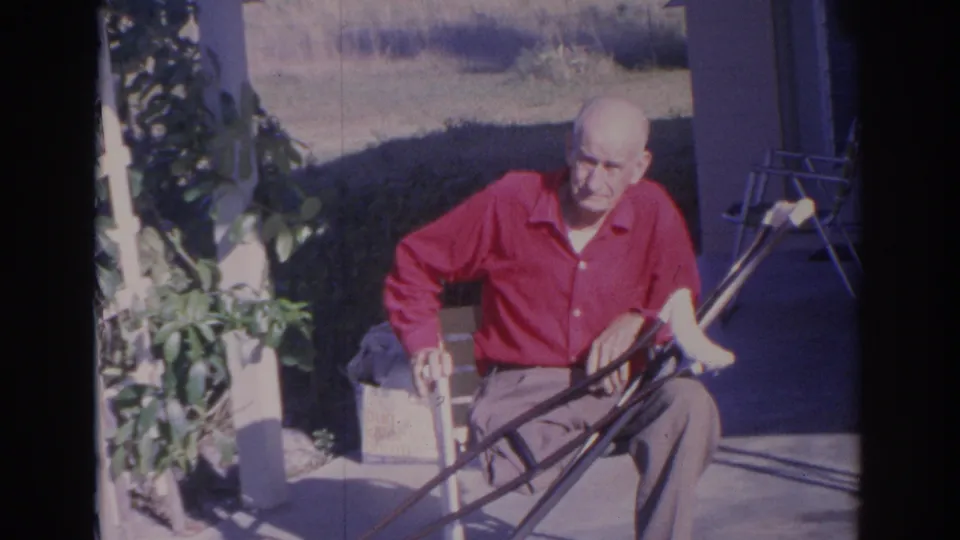An older man in a red shirt sits outside in Sarasota in 1969, framed by a doorway, trellis, patio, and garden plants.