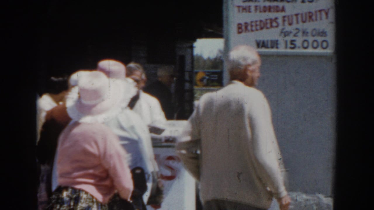 Older adults in hats stand outside a building near a Breeders Futurity sign.