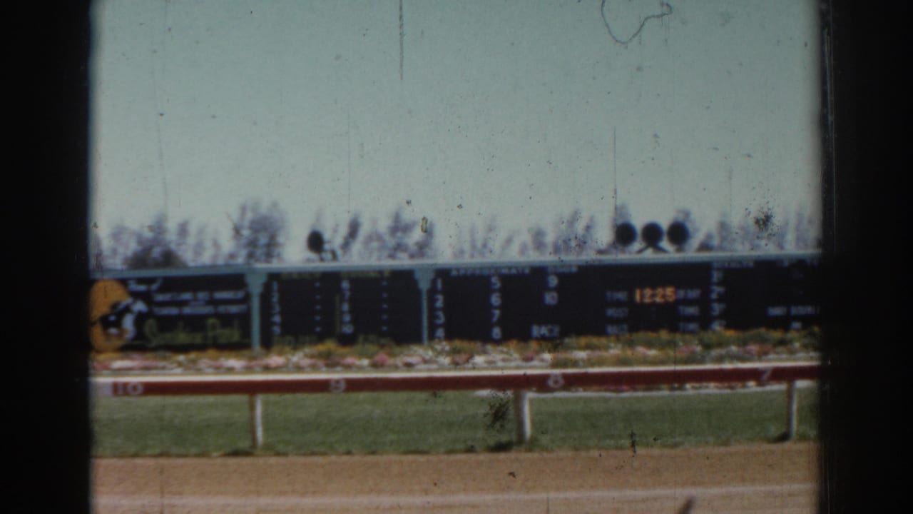 A handheld 8mm view of the Sarasota racetrack scoreboard, rail, grass, and dirt track.
