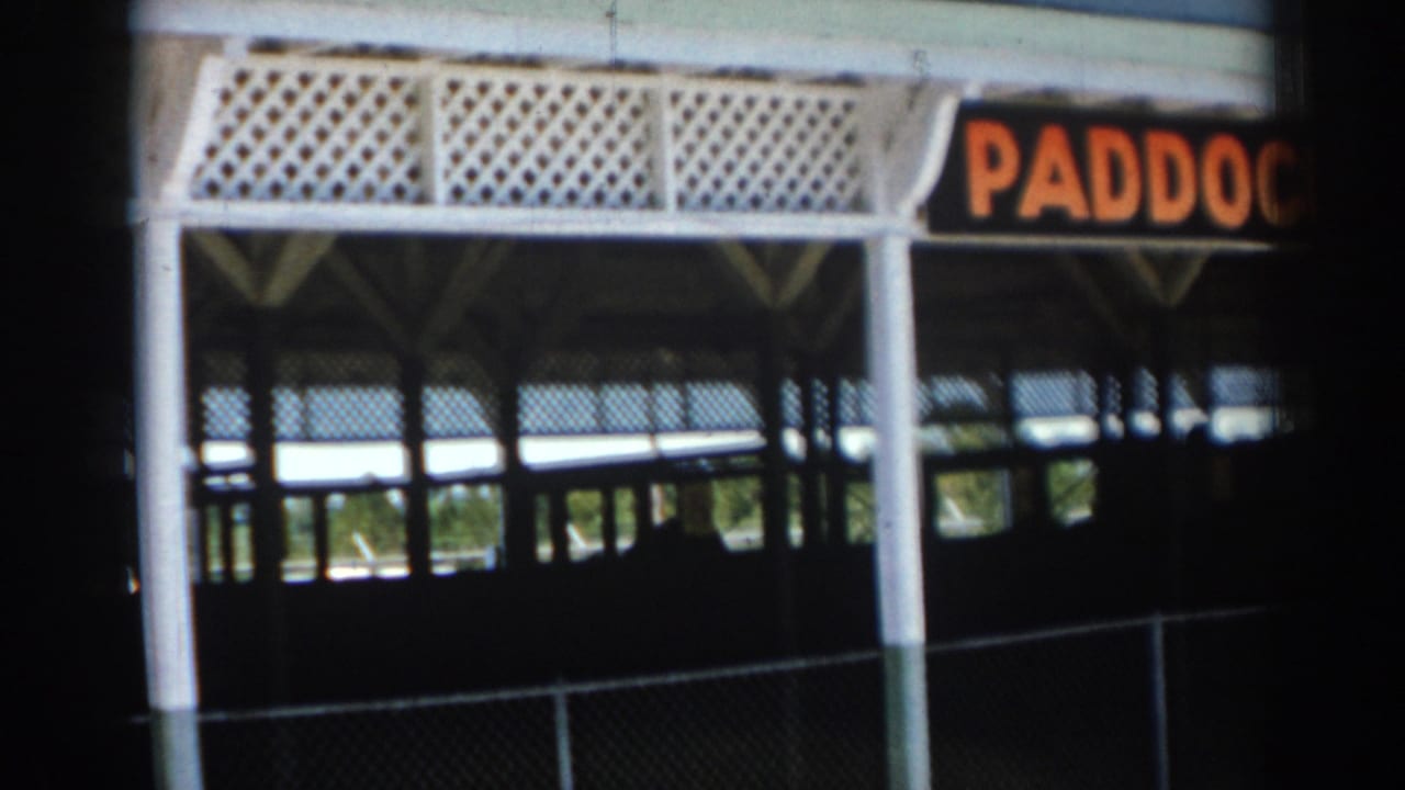 1969 Sarasota home-movie frame showing a white-railed paddock-area structure with a chain-link fence and shaded interior space.