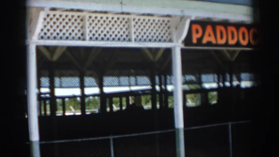 1969 Sarasota home-movie frame showing a white-railed paddock-area structure with a chain-link fence and shaded interior space.