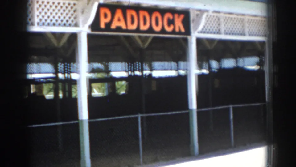 The Sarasota racetrack paddock sign fills the frame above a fenced, shaded walkway in 1969 8mm footage.