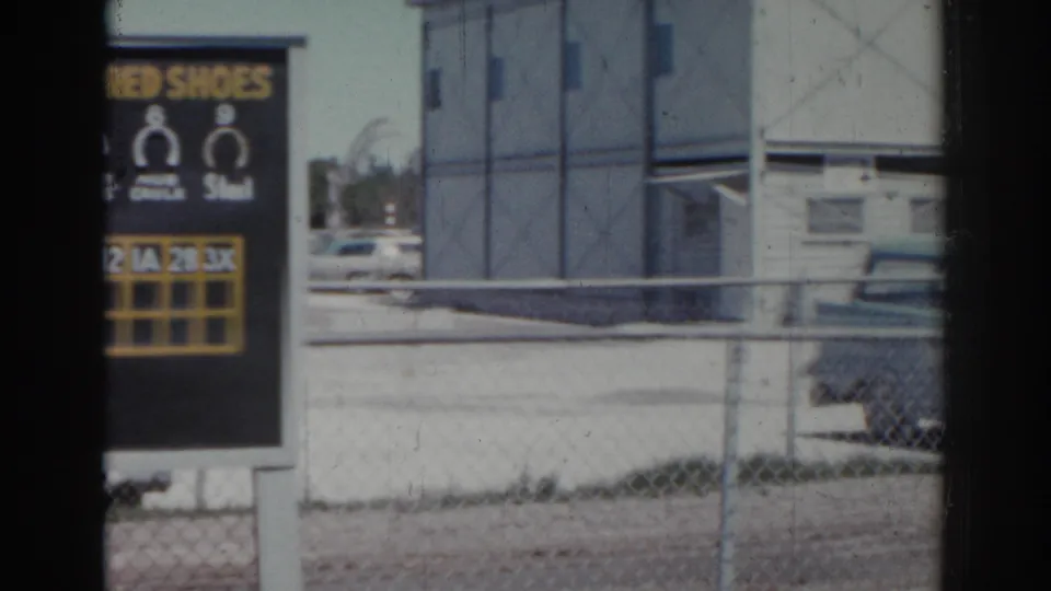 End of the pan shows the scoreboard edge near a chain-link fence, low trackside building, posts, and parked cars at Sunshine Park.
