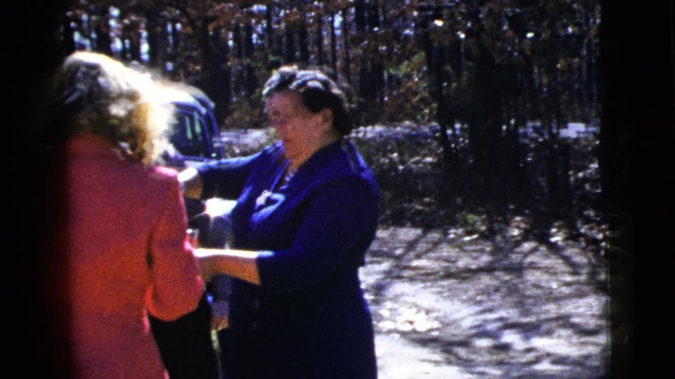 Frame from clip 124557771: In a color home movie outdoors, a person in dark blue holds a tray with small vessels and a bottle. A person in a coral-colored suit stands with their back partly toward the camera, then turns and smiles. Another person is partly visible behind them. A blue car, a fence and trees occupy the background. Dark vertical film edges frame the gathering. The short gesture repeats. No conversation has been assigned to these people.