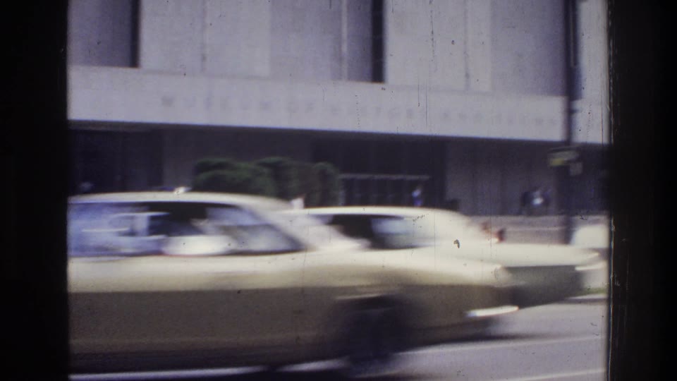 Frame from clip 121472189: In a color home movie, the camera looks through a dark-edged vehicle window across a road. A car and a red delivery truck pass in the foreground. The view moves along a pale building with an inscription reading Museum of History and Technology. Small people stand or walk near its front. More cars cross the image; near the end, white fountain sprays appear behind them. The short view repeats.